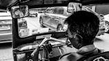 A black-and-white photograph captures the interior of a tuk-tuk, focusing on the driver as he navigates through a street filled with cars. The rearview mirror reflects the driver wearing glasses and a shirt. There are visible documents and license stickers on the dashboard.