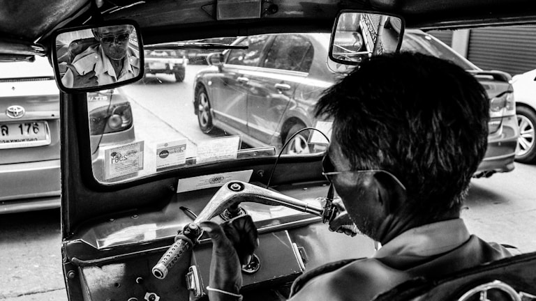 A black-and-white photograph captures the interior of a tuk-tuk, focusing on the driver as he navigates through a street filled with cars. The rearview mirror reflects the driver wearing glasses and a shirt. There are visible documents and license stickers on the dashboard.