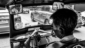 A black-and-white photograph captures the interior of a tuk-tuk, focusing on the driver as he navigates through a street filled with cars. The rearview mirror reflects the driver wearing glasses and a shirt. There are visible documents and license stickers on the dashboard.