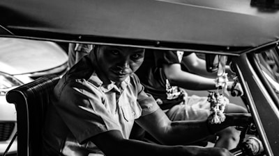 A black and white photograph of a person who appears to be driving a vehicle, likely a tricycle or small taxi. The person is wearing a collared shirt and looking directly at the camera with a serious expression. Another person is partially visible in the background, also inside the vehicle. The interior of the vehicle includes a steering mechanism and decorative hanging items.