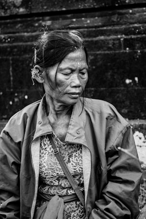 An elderly woman, seated and looking downward with a contemplative expression. She wears a lace top beneath a lightweight jacket and has a simple hairstyle with a flower tucked behind her ear. The background is blurred stone, giving an impression of an outdoor setting.