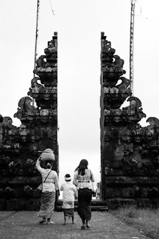 Three individuals are dressed in traditional attire, walking towards a large, intricately designed stone gate. One person is carrying a basket on their head. The scene is captured in black and white, emphasizing the historical and cultural elements of the architecture.