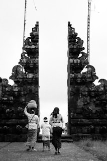 Three individuals are dressed in traditional attire, walking towards a large, intricately designed stone gate. One person is carrying a basket on their head. The scene is captured in black and white, emphasizing the historical and cultural elements of the architecture.