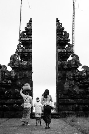 Three individuals are dressed in traditional attire, walking towards a large, intricately designed stone gate. One person is carrying a basket on their head. The scene is captured in black and white, emphasizing the historical and cultural elements of the architecture.