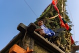 A young person stands on a decorated structure adorned with greenery and vibrant ribbons in red, blue, and green. Intricate carvings and ornate details can be observed on the stone or wooden parts of the structure.