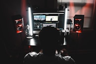 A person is sitting in front of a computer monitor displaying video editing software. The room is dimly lit with two tall lights flanking the monitor, casting a soft glow. High-quality studio speakers are positioned on both sides of the desk. The atmosphere suggests focused work or creativity.