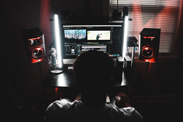 A person is sitting in front of a computer monitor displaying video editing software. The room is dimly lit with two tall lights flanking the monitor, casting a soft glow. High-quality studio speakers are positioned on both sides of the desk. The atmosphere suggests focused work or creativity.