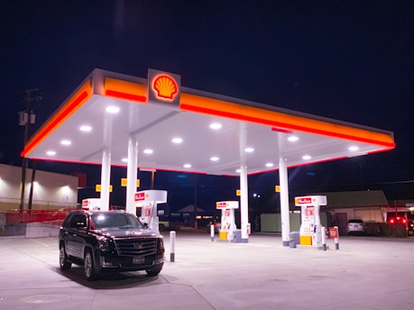 A well-lit gas station at night featuring a Shell canopy with illuminated signage. A black SUV is parked near the gas pumps, and the area is quiet with no visible people. The surrounding environment is dark, emphasizing the bright lights of the station.