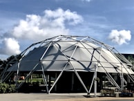 A geodesic dome structure made of metal framework and fabric, set against a backdrop of a bright blue sky with scattered white clouds. Lush green foliage surrounds the base of the dome, providing a contrast to the industrial design. Shadows from the framework are cast on the fabric, creating interesting patterns.