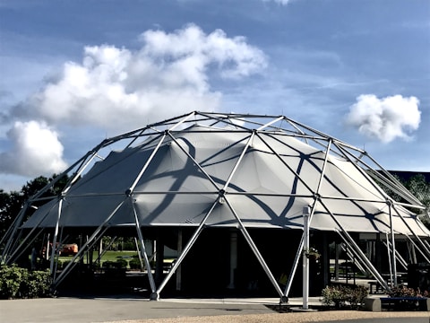 A geodesic dome structure made of metal framework and fabric, set against a backdrop of a bright blue sky with scattered white clouds. Lush green foliage surrounds the base of the dome, providing a contrast to the industrial design. Shadows from the framework are cast on the fabric, creating interesting patterns.