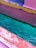 A close-up view of a boat's surface painted in vibrant colors, including pink, orange, yellow, and blue, with water seen close to the edge in a bright turquoise hue. The paint appears to be chipped and weathered with visible texture.