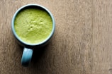 A blue mug filled with frothy green matcha latte placed on a textured wooden surface. The top of the latte appears creamy with small bubbles visible.