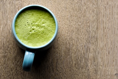 A blue mug filled with frothy green matcha latte placed on a textured wooden surface. The top of the latte appears creamy with small bubbles visible.