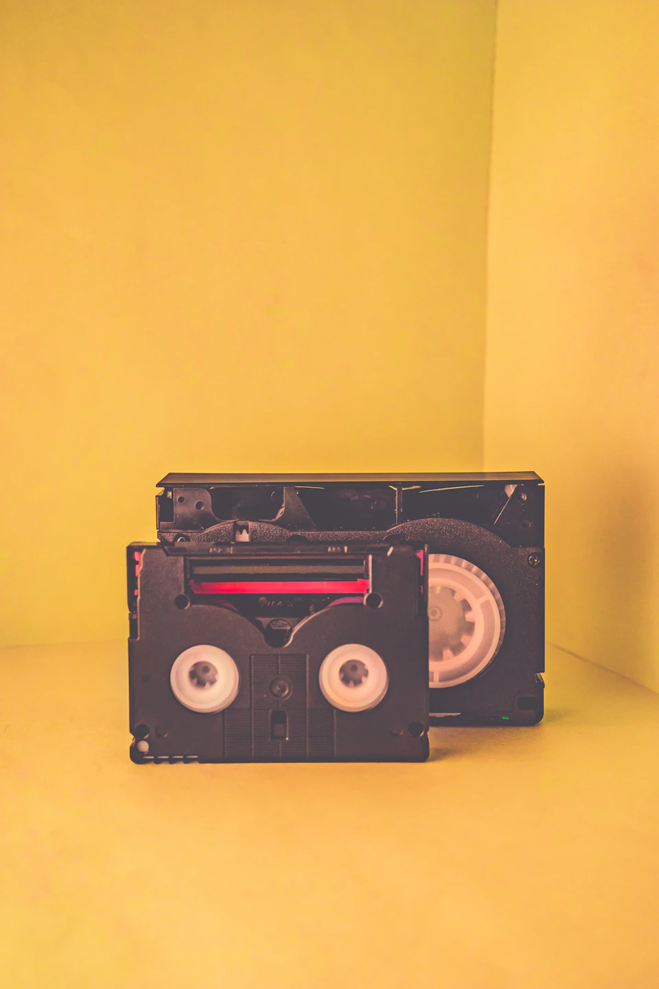 The image shows two black VHS tapes resting on a yellow background. The tape on the left is slightly larger than the one on the right, with the smaller tape positioned at an angle to the larger one. Both tapes appear to be in good condition and are facing towards the viewer.