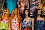 A row of four mannequins with long hair and distinct facial features, each wearing colorful and patterned summer dresses. The garments range from floral designs to geometric patterns, adding a variety of textures and colors. Behind them, a variety of other clothing items are displayed, including graphic t-shirts and additional patterned dresses.