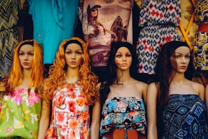 A row of four mannequins with long hair and distinct facial features, each wearing colorful and patterned summer dresses. The garments range from floral designs to geometric patterns, adding a variety of textures and colors. Behind them, a variety of other clothing items are displayed, including graphic t-shirts and additional patterned dresses.