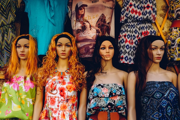 A row of four mannequins with long hair and distinct facial features, each wearing colorful and patterned summer dresses. The garments range from floral designs to geometric patterns, adding a variety of textures and colors. Behind them, a variety of other clothing items are displayed, including graphic t-shirts and additional patterned dresses.
