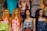 A row of four mannequins with long hair and distinct facial features, each wearing colorful and patterned summer dresses. The garments range from floral designs to geometric patterns, adding a variety of textures and colors. Behind them, a variety of other clothing items are displayed, including graphic t-shirts and additional patterned dresses.