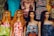 A row of four mannequins with long hair and distinct facial features, each wearing colorful and patterned summer dresses. The garments range from floral designs to geometric patterns, adding a variety of textures and colors. Behind them, a variety of other clothing items are displayed, including graphic t-shirts and additional patterned dresses.