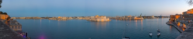 A panoramic view of a harbor with calm waters set against an expansive cityscape. Historic buildings with warm-toned facades line the water's edge, illuminated by the golden hues of sunset. Small boats are moored in the foreground while the distant skyline features various architectural structures including domes and towers.