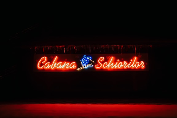 A vibrant neon sign displaying the words 'Cabana Schiorilor' with a blue skiing figure between the text. The sign is illuminated against a dark background, enhancing the glow of the red and blue lights.