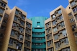 High-rise residential buildings with numerous windows and air conditioning units attached, displaying a combination of beige and teal colors under a clear blue sky.