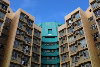 High-rise residential buildings with numerous windows and air conditioning units attached, displaying a combination of beige and teal colors under a clear blue sky.