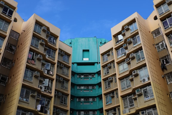 High-rise residential buildings with numerous windows and air conditioning units attached, displaying a combination of beige and teal colors under a clear blue sky.