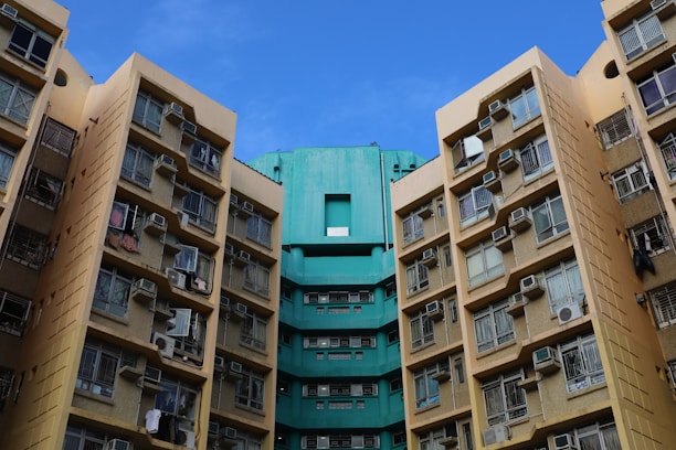 High-rise residential buildings with numerous windows and air conditioning units attached, displaying a combination of beige and teal colors under a clear blue sky.