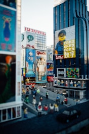 A bustling cityscape constructed entirely with LEGO bricks, featuring tall buildings with colorful advertisements and signs. The scene includes a variety of small LEGO figures depicting people walking and gathering in a street area. The buildings are detailed, with one having a prominent LEGO character on a billboard.