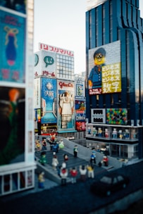 A bustling cityscape constructed entirely with LEGO bricks, featuring tall buildings with colorful advertisements and signs. The scene includes a variety of small LEGO figures depicting people walking and gathering in a street area. The buildings are detailed, with one having a prominent LEGO character on a billboard.