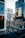 A bustling cityscape constructed entirely with LEGO bricks, featuring tall buildings with colorful advertisements and signs. The scene includes a variety of small LEGO figures depicting people walking and gathering in a street area. The buildings are detailed, with one having a prominent LEGO character on a billboard.