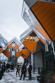 A group of people stand under a series of uniquely designed cube-shaped houses, characterized by their orange facades and geometric structures. The sky is overcast, and the scene has a modern, architectural focus.