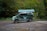 A van with a green, nature-themed wrap is parked on a dirt surface with puddles. Behind it is a shelter with a green roof, against a backdrop of lush, green forest.