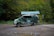 A van with a green, nature-themed wrap is parked on a dirt surface with puddles. Behind it is a shelter with a green roof, against a backdrop of lush, green forest.