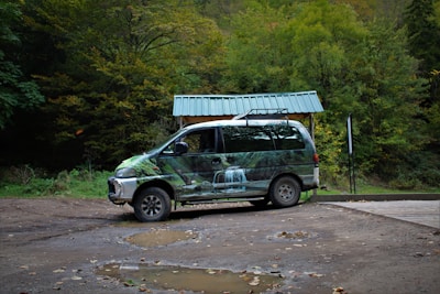 A van with a green, nature-themed wrap is parked on a dirt surface with puddles. Behind it is a shelter with a green roof, against a backdrop of lush, green forest.