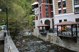 A serene stream flows alongside a multi-story brick and stucco building with balconies. The building features arched openings and is flanked by a path lined with trees and a wooden railing. The area includes decorative gazebos with wooden beams and a colorful play structure visible nearby. Lush greenery surrounds the setting, creating a peaceful and inviting atmosphere.