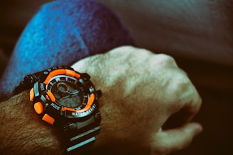 A close-up view of an analog-digital wristwatch with a sporty design on a person's wrist. The watch features bold orange elements on the bezel and buttons, contrasting with a black strap and casing. The wearer is dressed in blue denim, and the image has a subdued, vintage color tone.