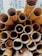 A collection of yellow cylindrical tubes is stacked closely together, creating a pattern of overlapping circles. The tubes appear to be weathered and vary in size, with some showing signs of wear and paint peeling. Buildings can be seen in the blurred background, suggesting an urban setting.