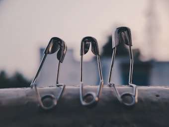 Three metallic safety pins are positioned upright on a surface. They are in a slightly hunched configuration, with strong focus on the glossy texture and reflection of light on the metal. The background is softly blurred, hinting at an urban setting with neutral colors.