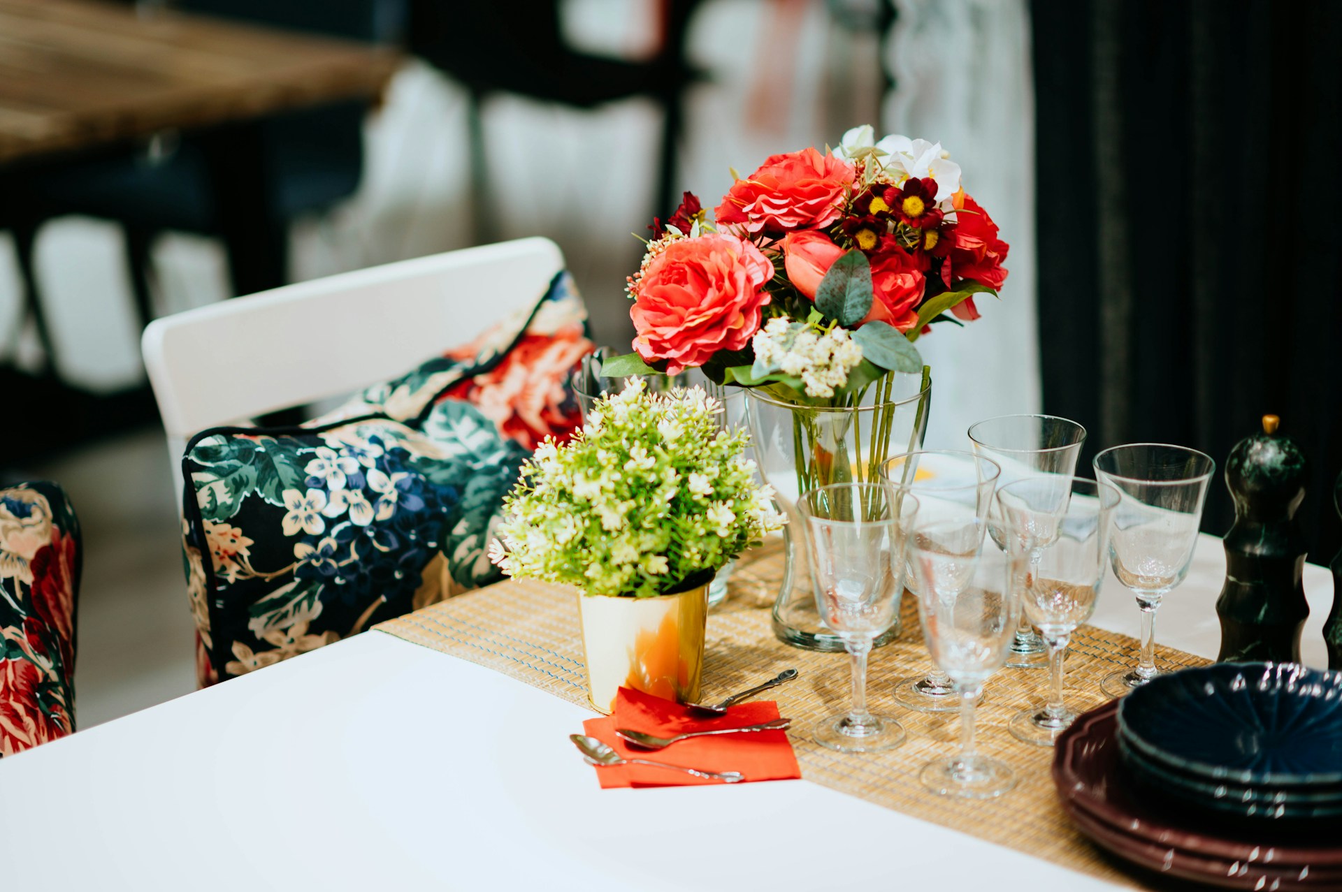 A table setting includes a floral-themed arrangement with a variety of flowers in red, white, and yellow placed in a glass vase. Adjacent is a smaller pot of green foliage. The table is set with several wine glasses, silverware, red napkins, and black plates neatly stacked. The background features chairs with floral-print cushions and other dining tables and chairs.