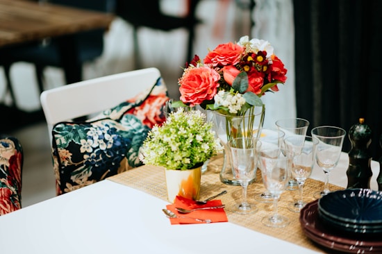 A table setting includes a floral-themed arrangement with a variety of flowers in red, white, and yellow placed in a glass vase. Adjacent is a smaller pot of green foliage. The table is set with several wine glasses, silverware, red napkins, and black plates neatly stacked. The background features chairs with floral-print cushions and other dining tables and chairs.