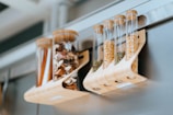 Transparent cylindrical containers hold various dried goods, such as mushrooms, seeds, and spices. They are supported by a wooden rack and attached to a metallic surface, likely a refrigerator. The setting appears modern and organized.