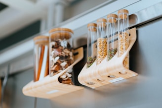 Transparent cylindrical containers hold various dried goods, such as mushrooms, seeds, and spices. They are supported by a wooden rack and attached to a metallic surface, likely a refrigerator. The setting appears modern and organized.