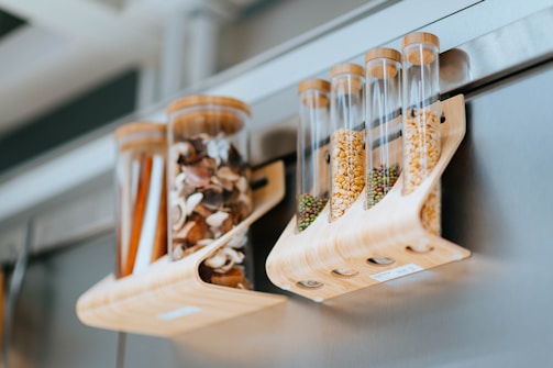 Transparent cylindrical containers hold various dried goods, such as mushrooms, seeds, and spices. They are supported by a wooden rack and attached to a metallic surface, likely a refrigerator. The setting appears modern and organized.
