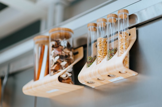 Transparent cylindrical containers hold various dried goods, such as mushrooms, seeds, and spices. They are supported by a wooden rack and attached to a metallic surface, likely a refrigerator. The setting appears modern and organized.