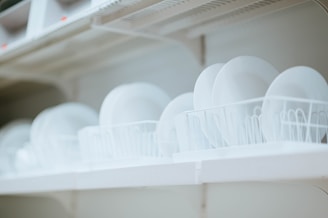 Stacks of clean white plates are neatly arranged on a shelf, held upright by wire dish racks. The scene conveys a sense of order and cleanliness, with a focus on the simplicity of the dishware.