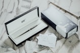 An elegant pen encased in a black and white box is opened to reveal its contents. Next to the box sits an accompanying blue and white outer packaging featuring drawings. A card with hand-written notes lies nearby on a textured surface, adding a personalized touch.