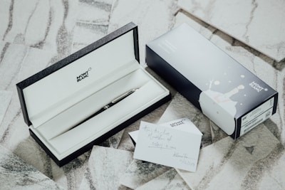 An elegant pen encased in a black and white box is opened to reveal its contents. Next to the box sits an accompanying blue and white outer packaging featuring drawings. A card with hand-written notes lies nearby on a textured surface, adding a personalized touch.