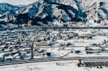 A sprawling town covered in snow lies at the base of rugged, forested mountains. Numerous buildings, both tall and short, are scattered throughout the area, while roads crisscross the urban landscape. The mountainous backdrop is heavily snow-laden, with dark trees creating a striking contrast. Several structures stand prominently near the mountains, suggesting a mix of residential and commercial use.